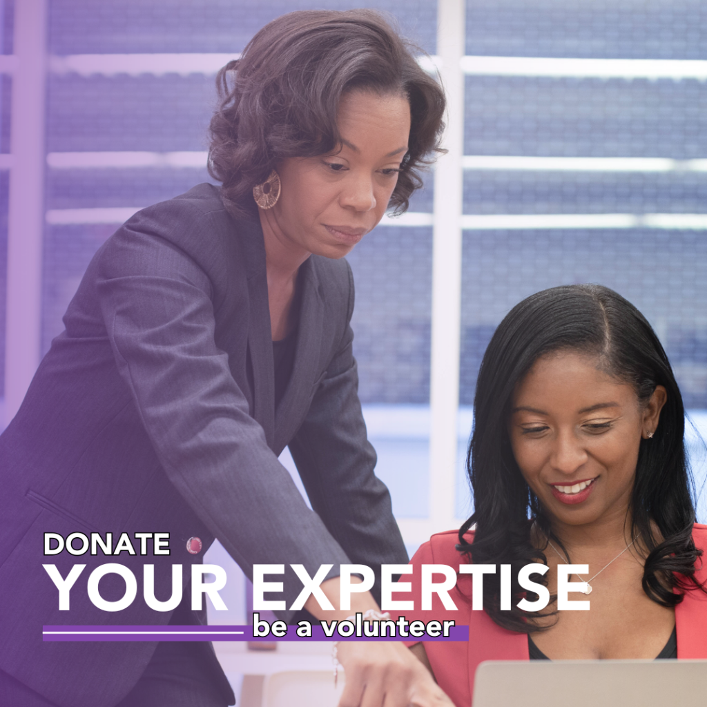 middle-aged African-American woman in dark suit pointing at a laptop of a young African-American woman sitting in a chair wearing orange shirt